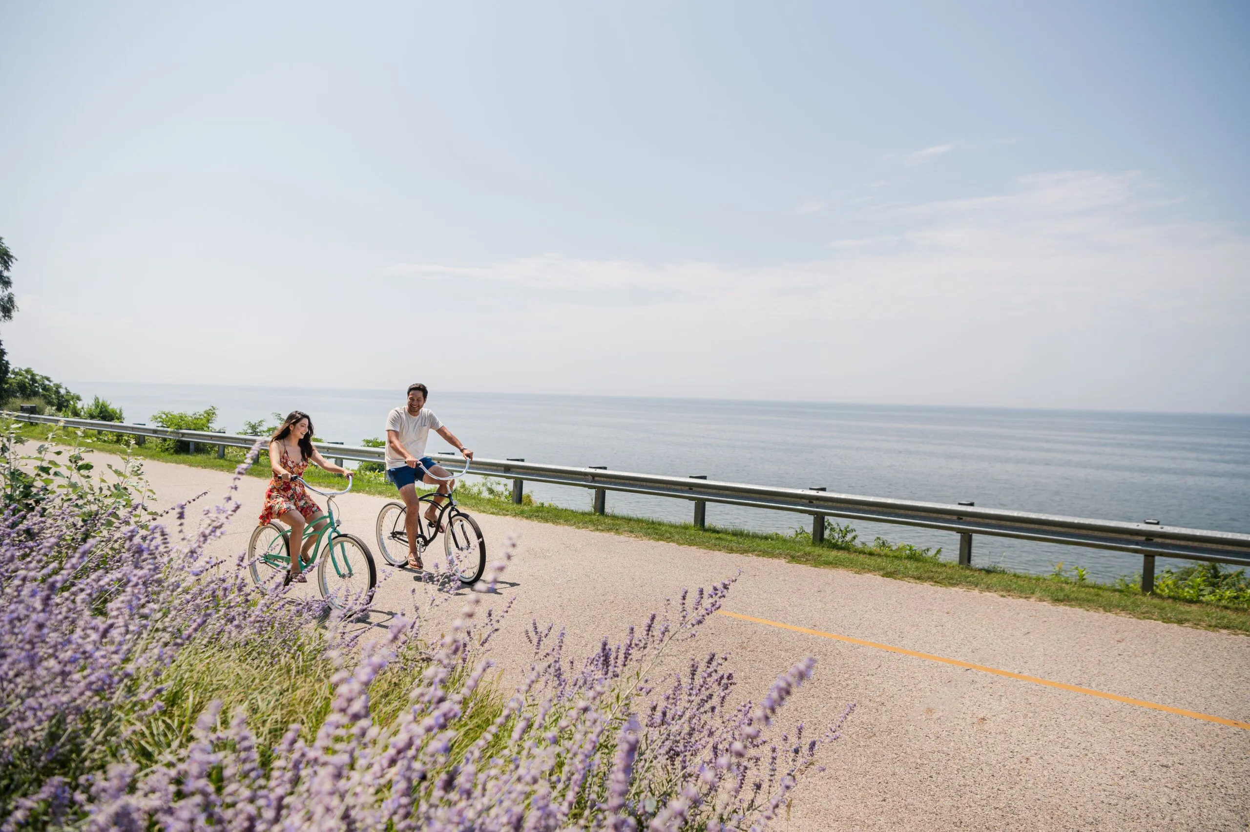 A couple smiling at each other and biking on a road near the water.