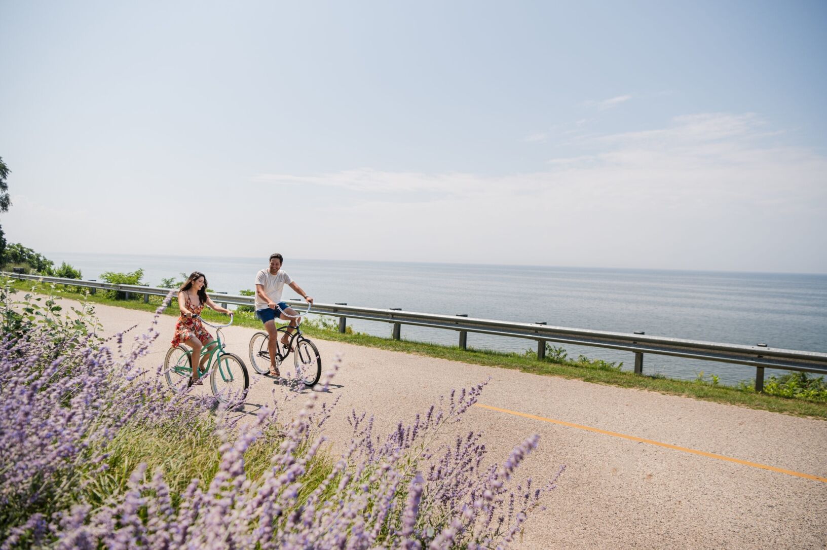 A couple smiling at each other and biking on a road near the water.
