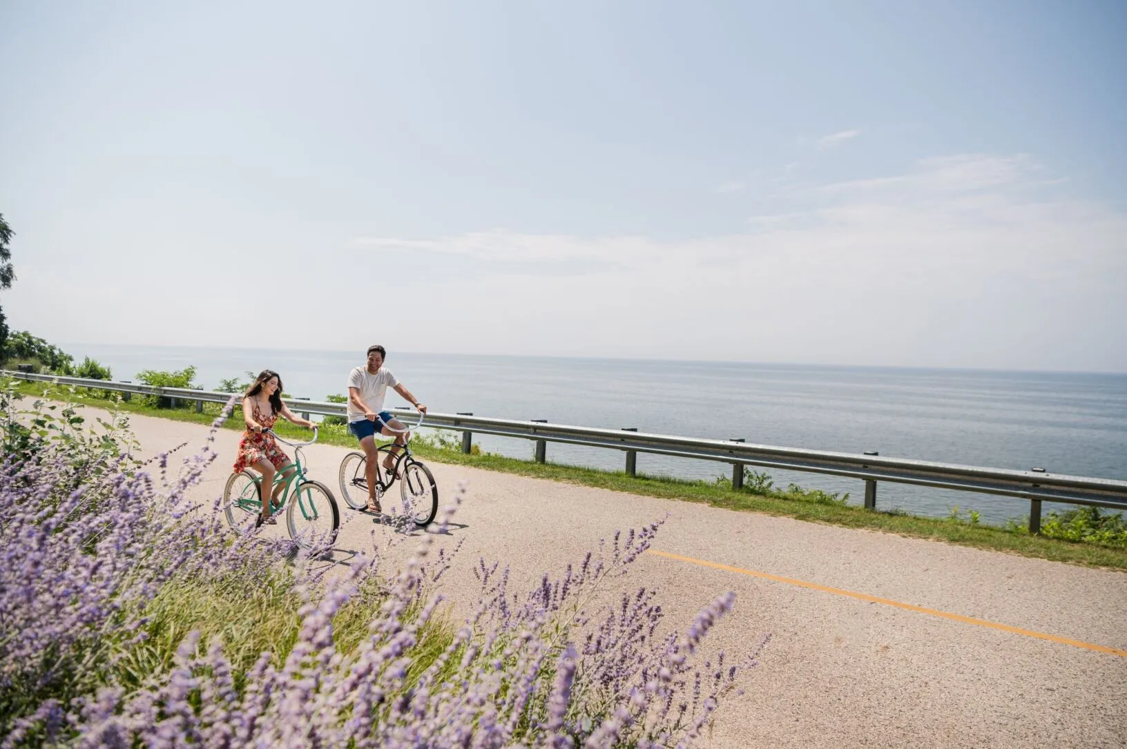 A couple smiling at each other and biking on a road near the water.