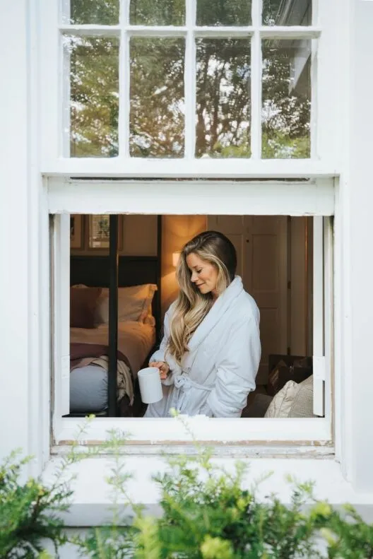 A woman in a robe holds a mug while looking outside a window.