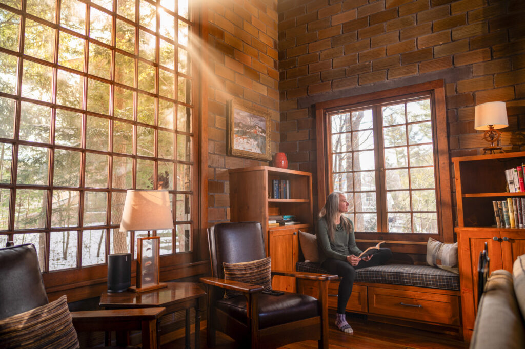 A relaxed woman with a book looks outside from a bench in a cozy rental home.