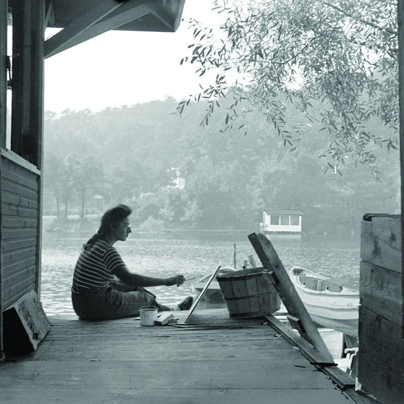 A black and white photo of an artist painting on the river.