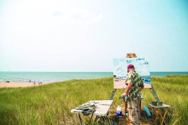 A man, set up with his easel in the grass, paints the lake.