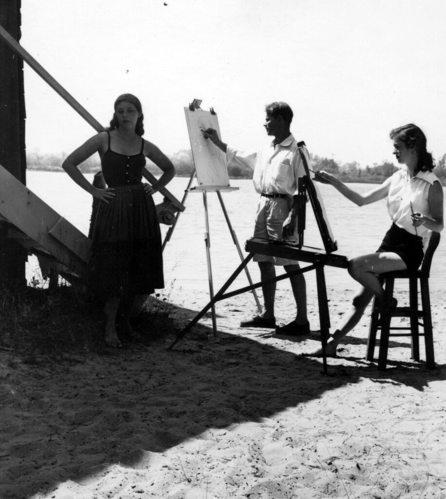 A black and white photo of two artists drawing a portrait of a woman outside.