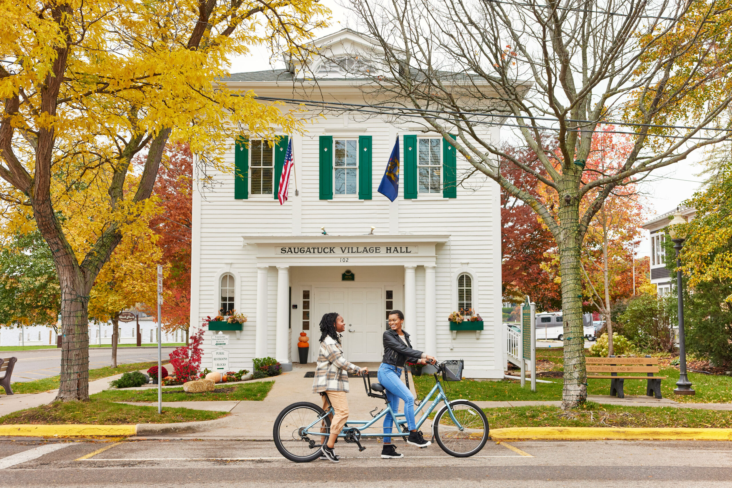 Two girls smile at each other as they ride bikes in front of the Saugatuck Village Hall. Golden fall colors and trees are in the background.