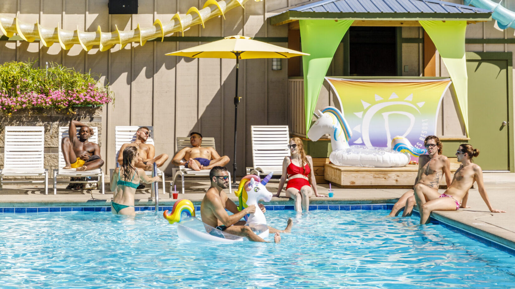 A group of people playing in a pool at The Dunes Resort. Some people are relaxing on the poolside while a man rides a float.
