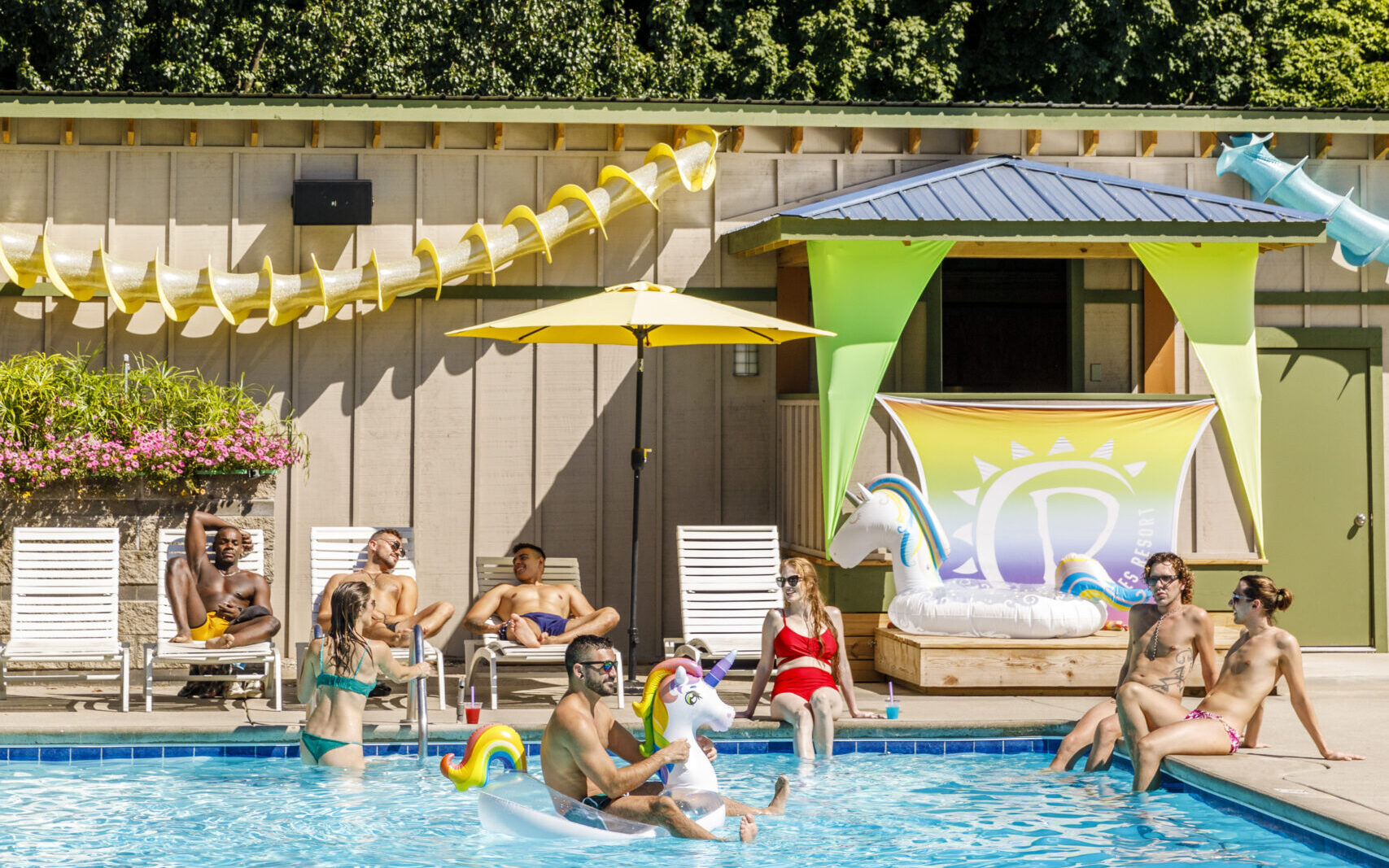 A group of people playing in a pool at The Dunes Resort. Some people are relaxing on the poolside while a man rides a float.