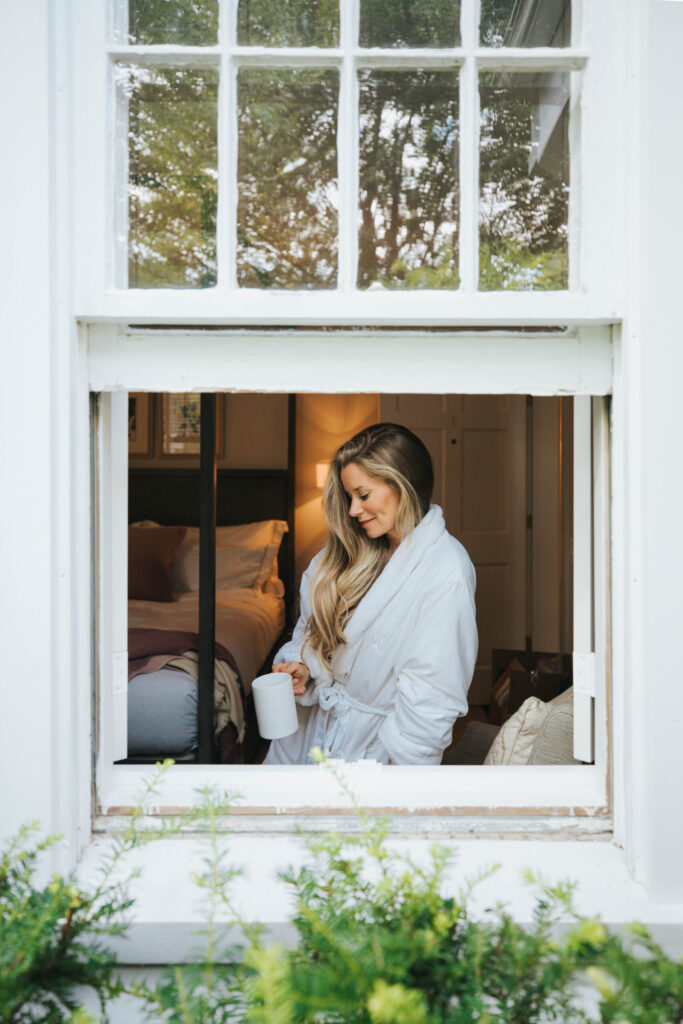 A woman in a robe holds a mug while looking outside a window.