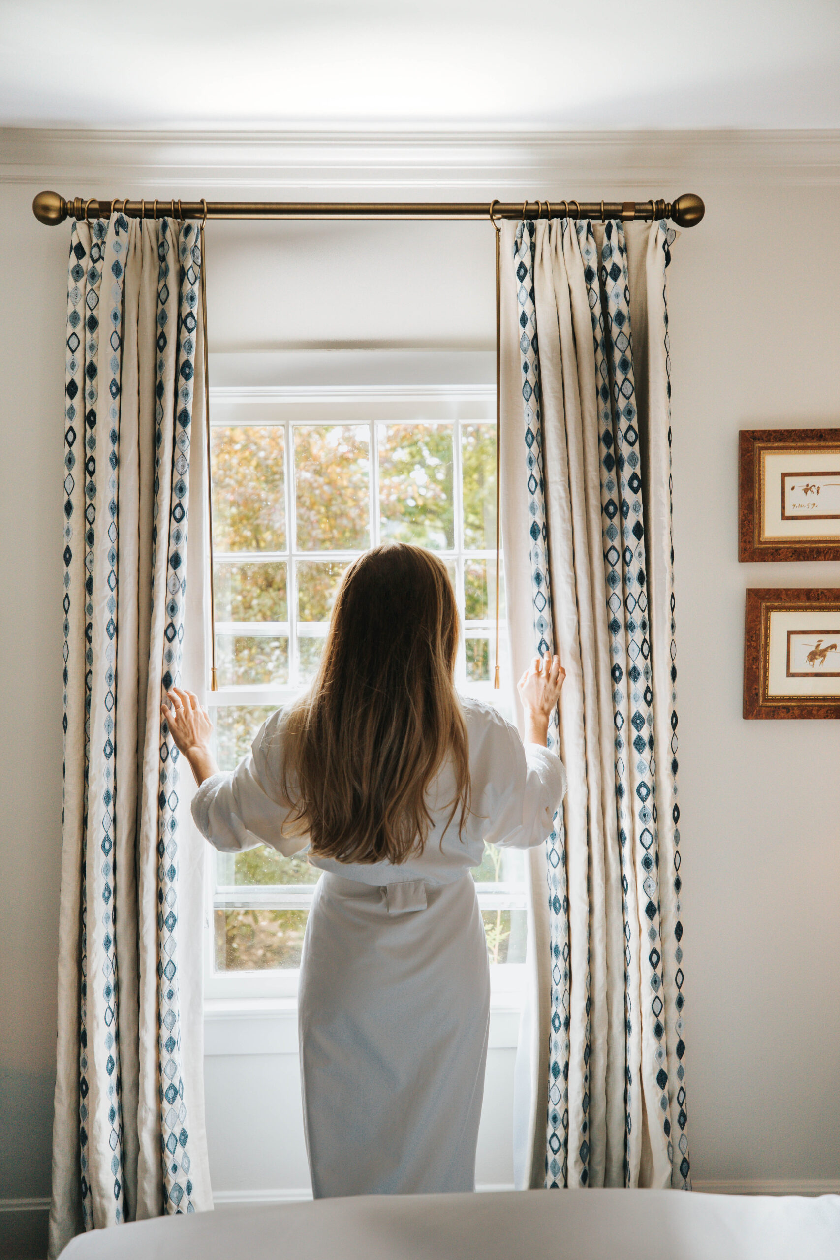 A woman in a robe draws the curtains to look outside a window.
